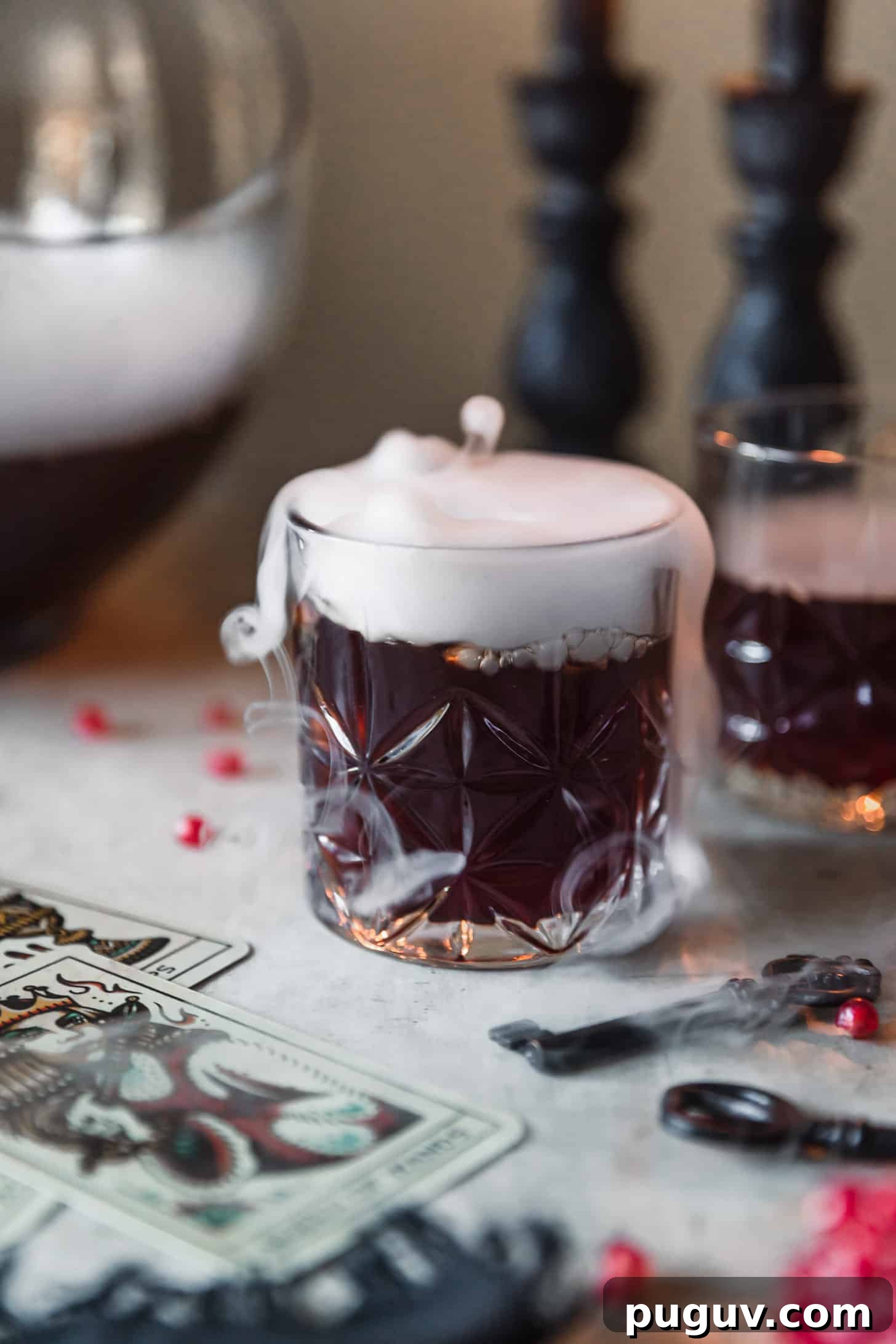 A glass of witches brew cocktail punch with dry ice on a grey counter next to a punch bowl, black candles, tarot cards, and pomegranate arils.