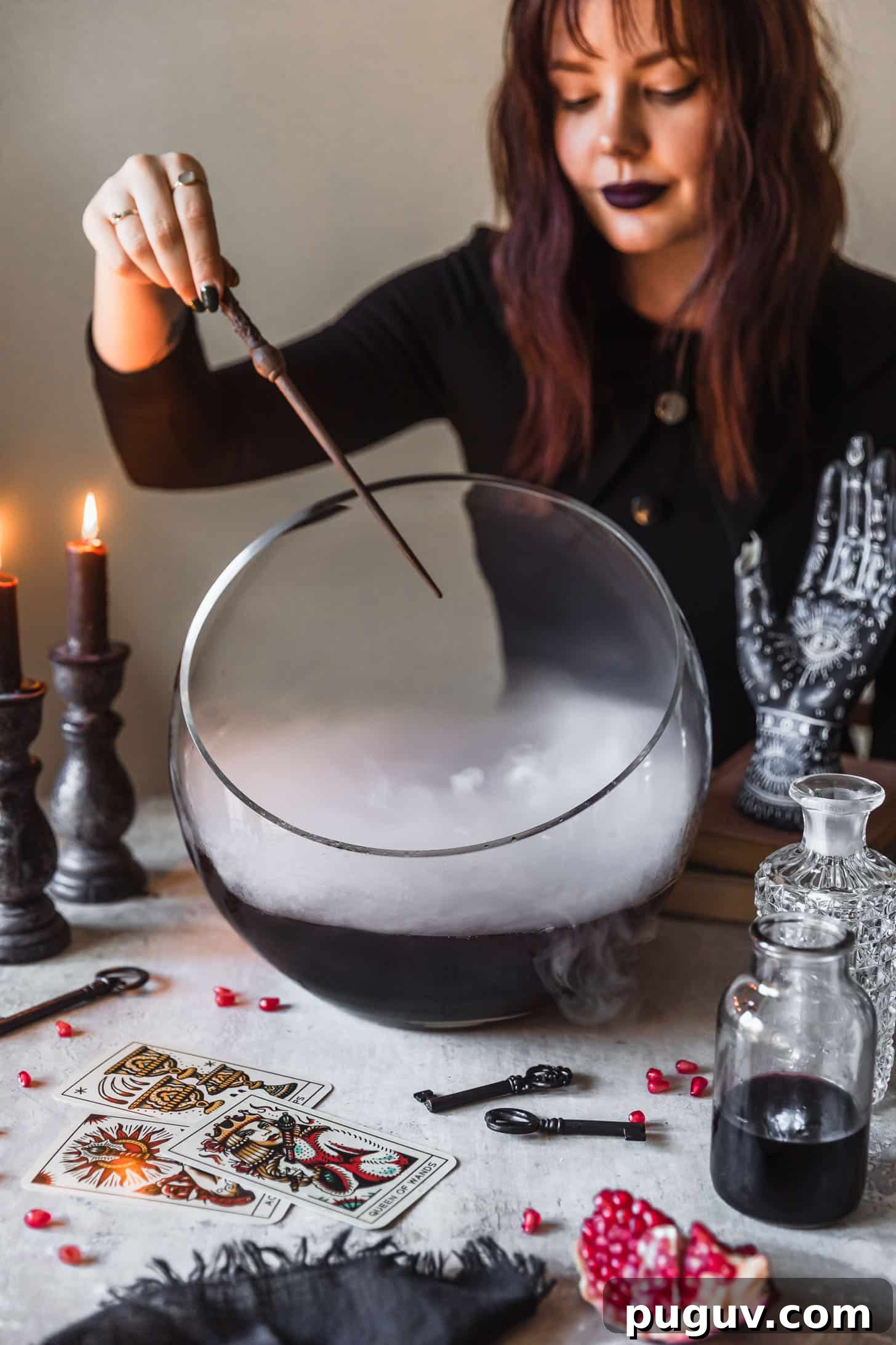 A woman in a black dress waving a wand over a bowl of witches brew cocktail punch next to potion bottles, vintage keys, and tarot cards on a grey background.