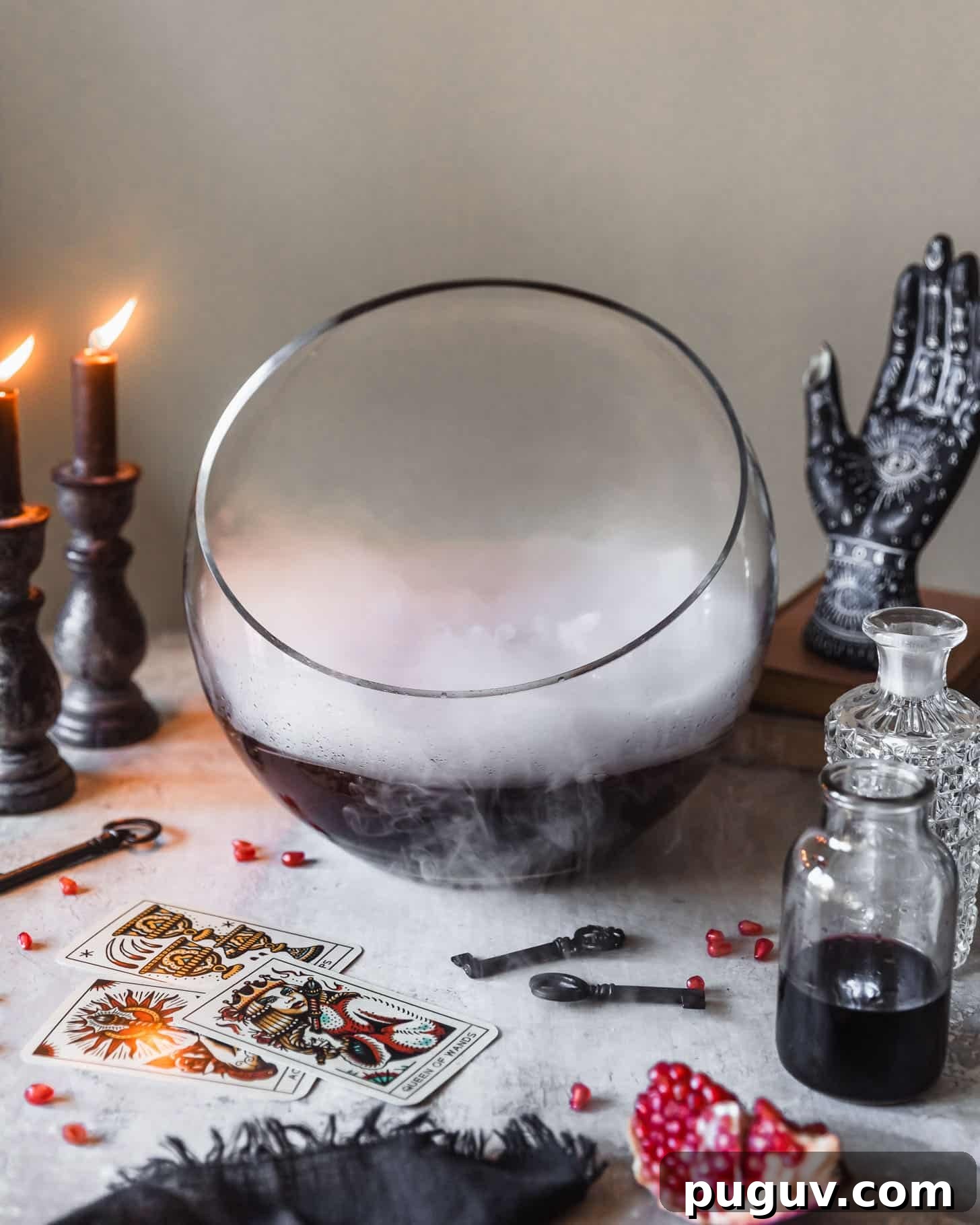 A bowl with a purple Halloween drink on a white counter next to lit black candles, bottles of alcohol, Halloween decorations, and tarot cards.