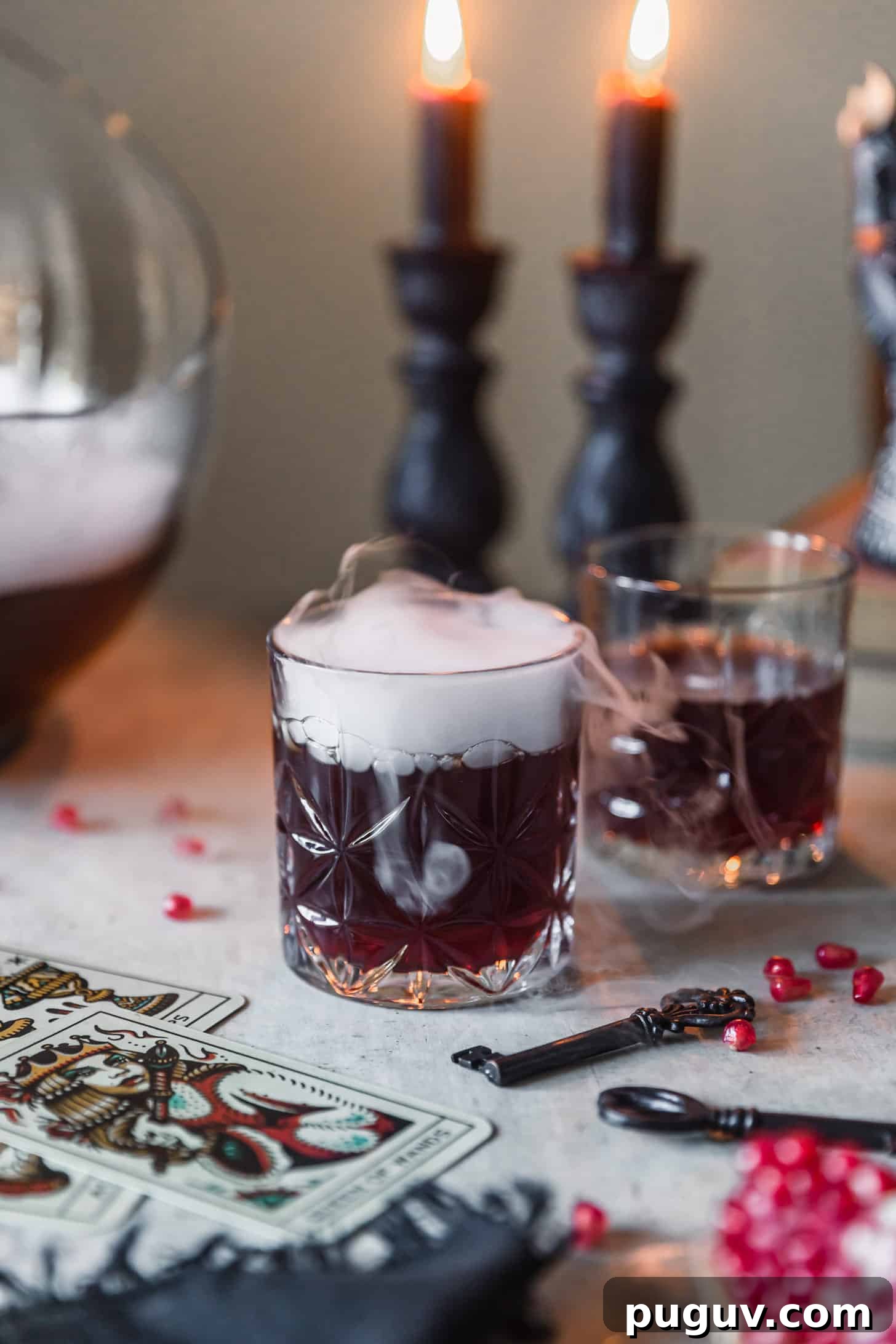 Two witches brew cocktails on a grey table next to black candles, pomegranate arils, vintage keys, and cards.