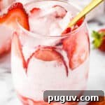 Side close up shot of a glass tumbler with strawberry mousse and sliced strawberries with a gold spoon on a marble table.