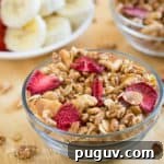 Side view of granola and strawberries in a clear bowl on a wooden surface with more granola and sliced fruits in the background.