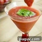 Side angled shot of a frozen strawberry basil margarita garnishe with basil leaves and a strawberry on the rim of the margarita glass with a red bottom with a bowl of strawberries in the background.