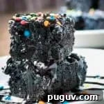 Side shot of two black bean brownies stacked on each other spinkled with powdered sugar, chocolate syrup, and rainbow sprinkles on a white plate.