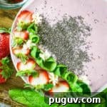 Overhead shot of pink smoothie bowl garnished with flax seeds, sliced strawberries, and mint rolls on a wooden surface with more mint and strawberries on the side.