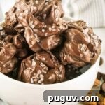 Side shot of multiple chocolate nut clusters with sea salt in a white bowl with almonds and chocolate chips sprinkled around.