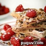 Side shot of multiple waffle pieces stacked on each other with raspberries and chocolate rice krispies and topped with powdered sugar on a red and white plate.