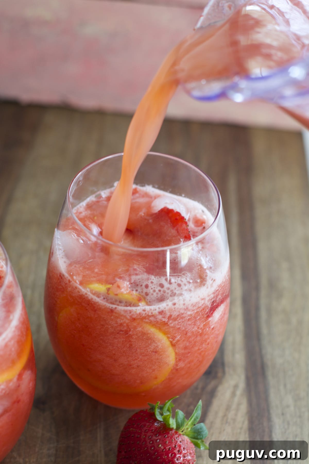 a pitcher of frozen lemonade with strawberries pouring into a glass with sliced lemons and berries