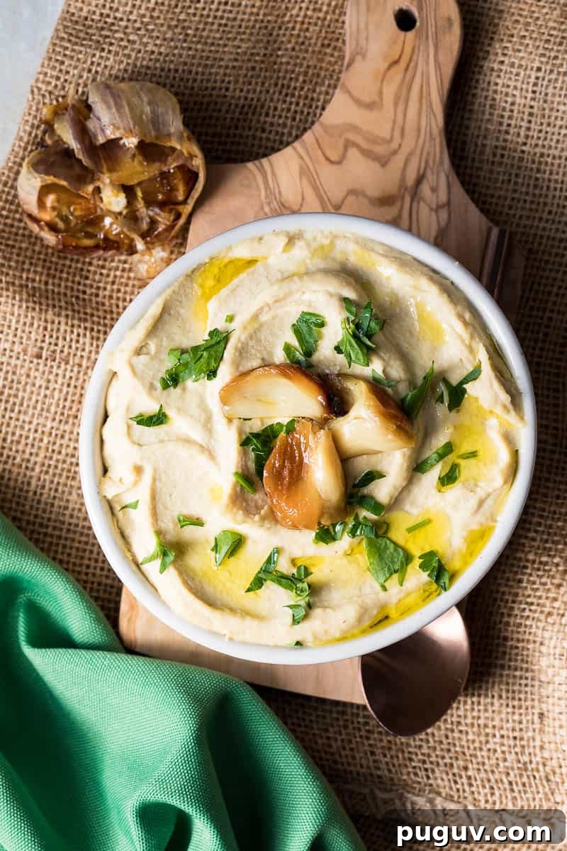 Creamy Roasted Garlic Hummus Recipe for Smooth, Flavorful Dip 2 bowl of roasted garlic hummus on cutting board sitting on burlap next to green napkin and caramelized garlic head.