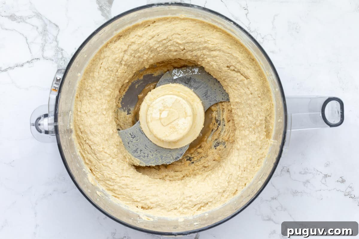 Creamy Roasted Garlic Hummus Recipe for Smooth, Flavorful Dip 5 chickpeas, tahini, and roasted garlic being processed in bowl of food processor.