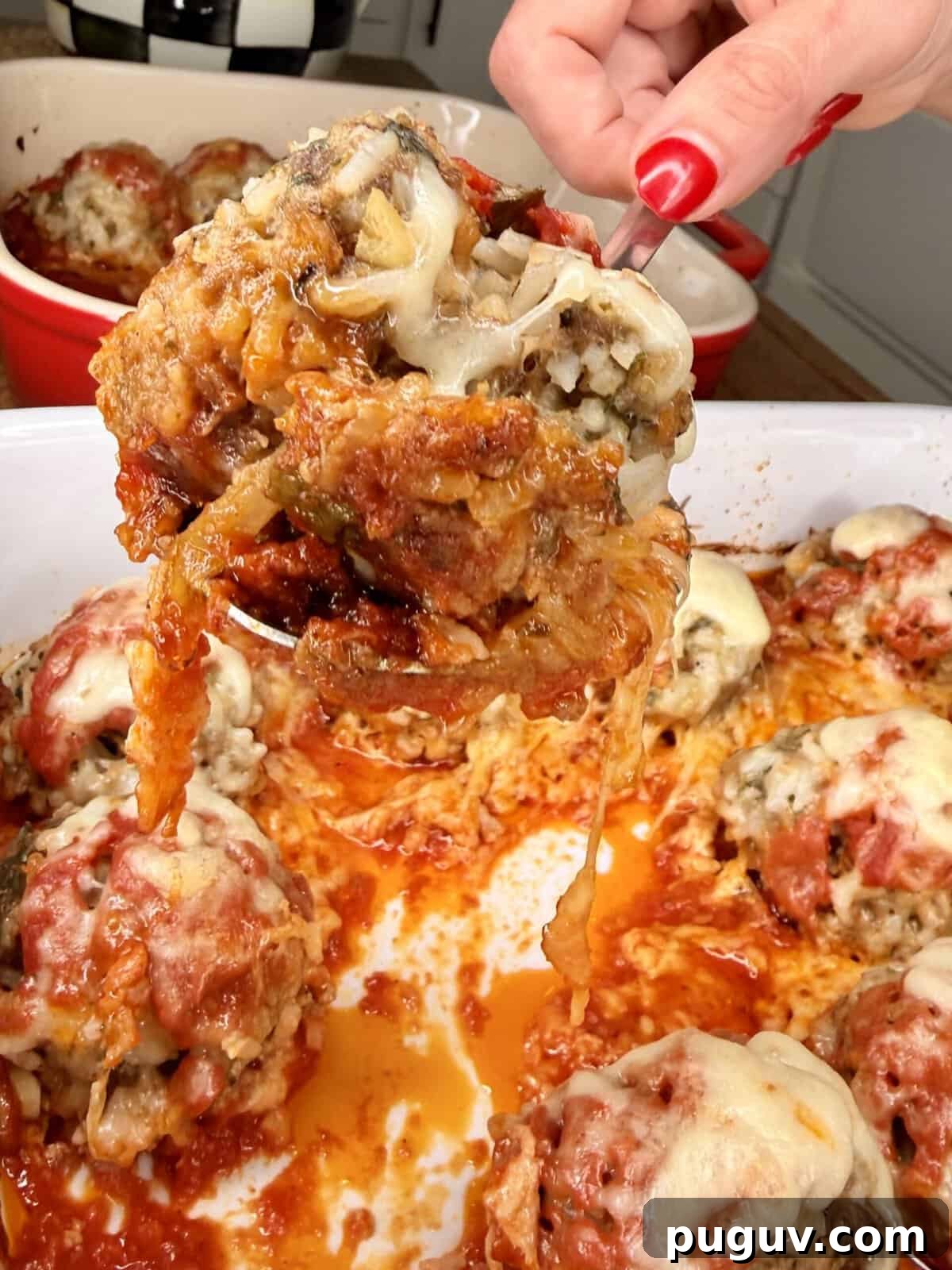 Italian-Style Baked Rice and Meatball Casserole 11 A hand lifts a cheesy, saucy stuffed meatball from a white baking dish filled with similar meatballs topped with melted cheese and tomato sauce.