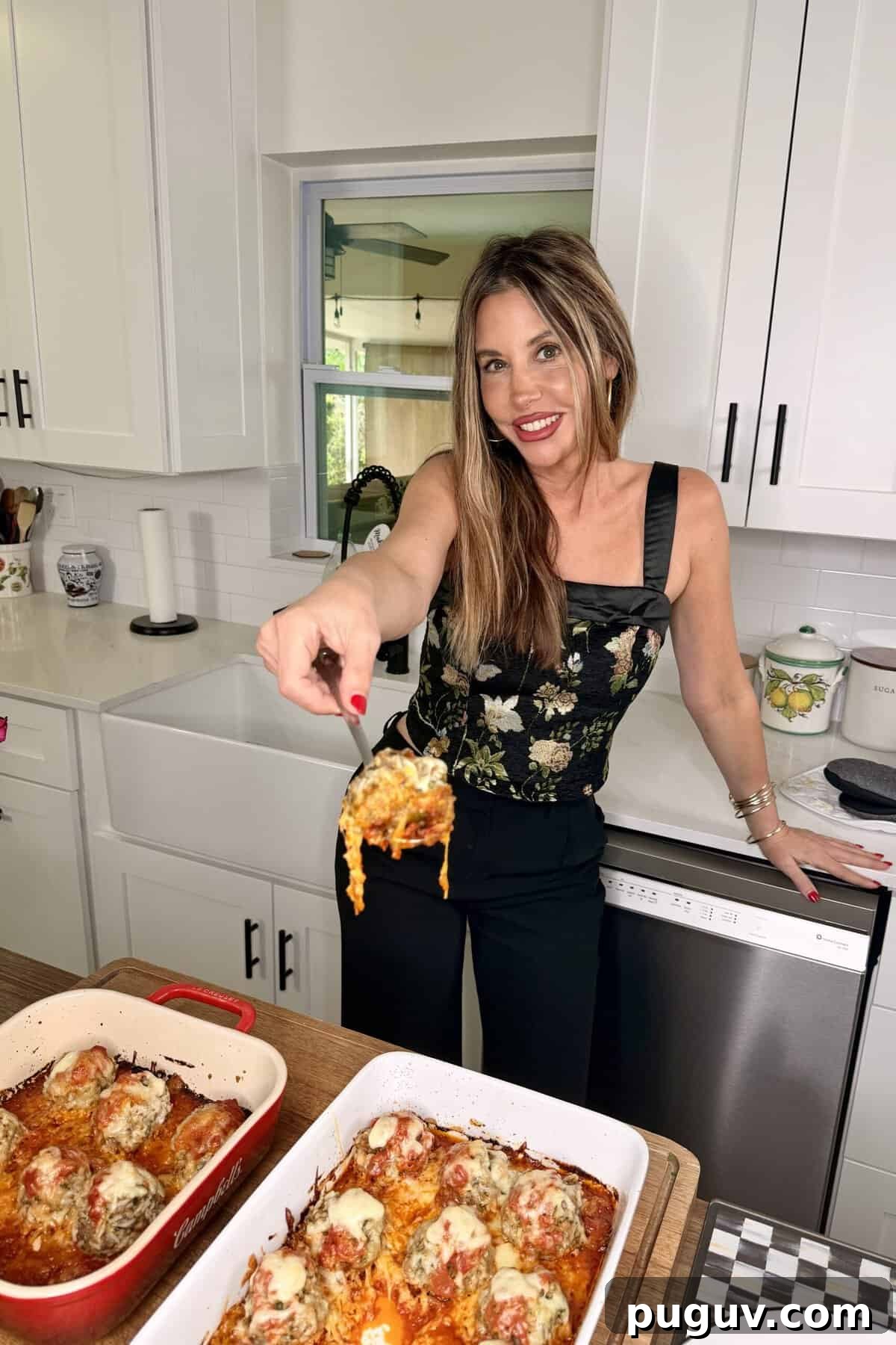 Italian-Style Baked Rice and Meatball Casserole 13 A woman in a black floral top smiles in a kitchen, holding up a serving of cheesy baked meatballs from a casserole dish on a wooden counter. Another dish of meatballs is also visible.