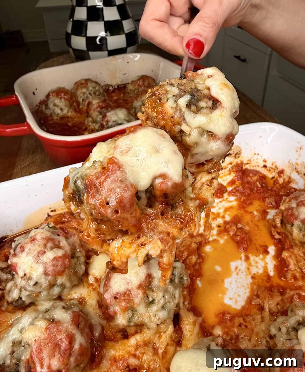 Italian-Style Baked Rice and Meatball Casserole 15 A hand with red nails lifts a cheesy, baked meatball covered in marinara sauce from a casserole dish filled with similar meatballs. A second dish with more meatballs sits in the background.