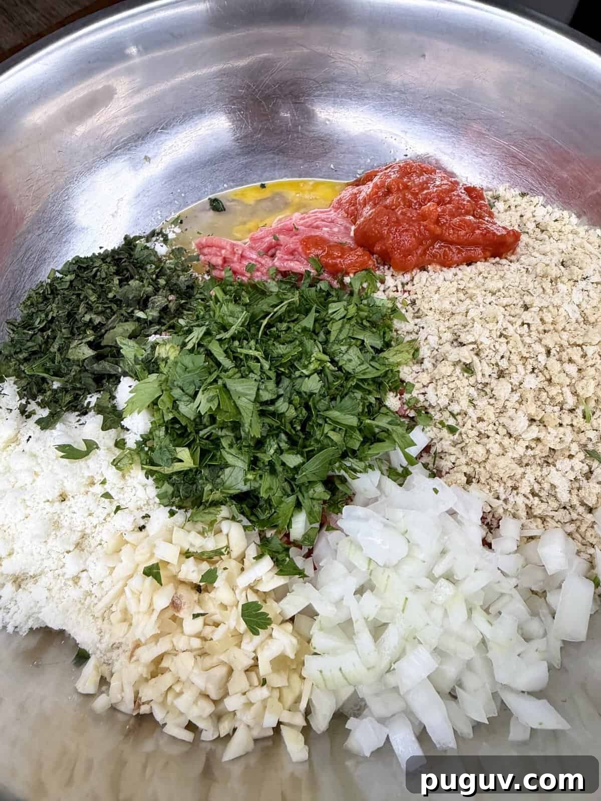 Italian-Style Baked Rice and Meatball Casserole 3 A metal bowl contains chopped parsley, chopped onion, minced garlic, dried herbs, tomato sauce, breadcrumbs, shredded cheese, egg, and ground meat, all arranged in separate piles, ready to be mixed.