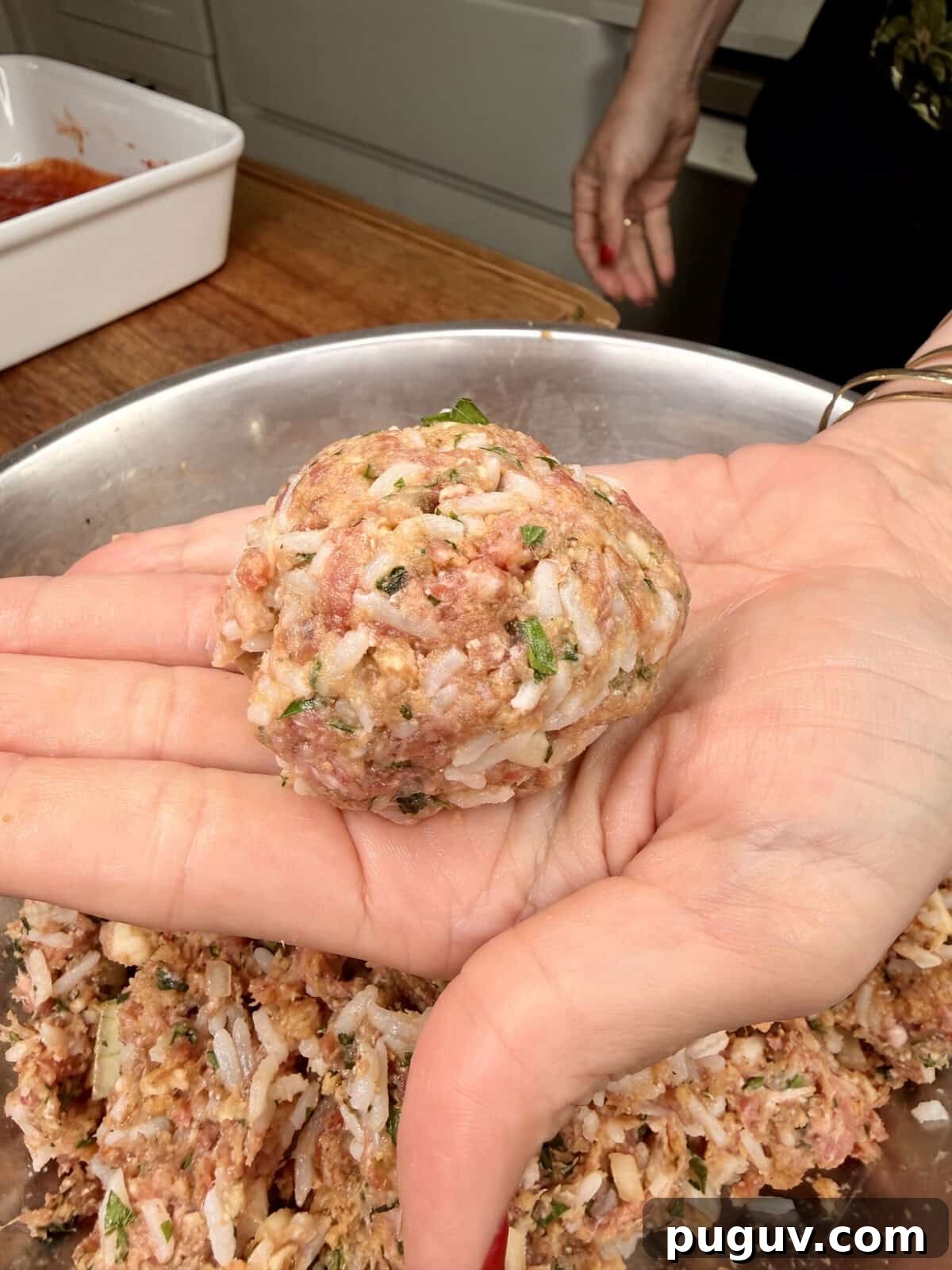 Italian-Style Baked Rice and Meatball Casserole 6 A close-up of a hand holding a large, uncooked meatball mixture with rice, herbs, and ground meat over a metal bowl filled with more mixture. Another hand rests on a wooden countertop in the background.