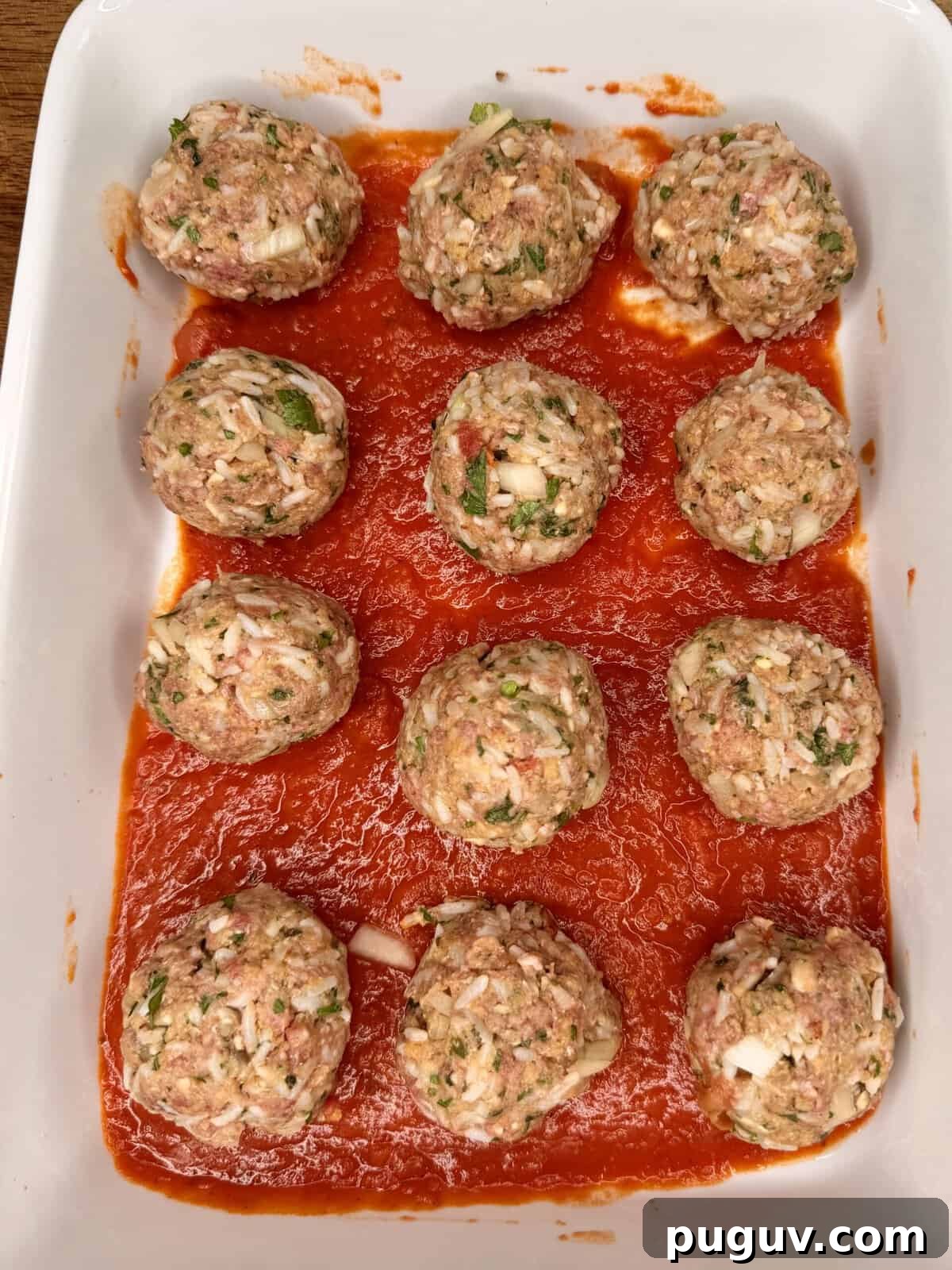 Italian-Style Baked Rice and Meatball Casserole 7 A white baking dish with twelve raw meatballs, mixed with chopped onions and herbs, arranged on a layer of tomato sauce.