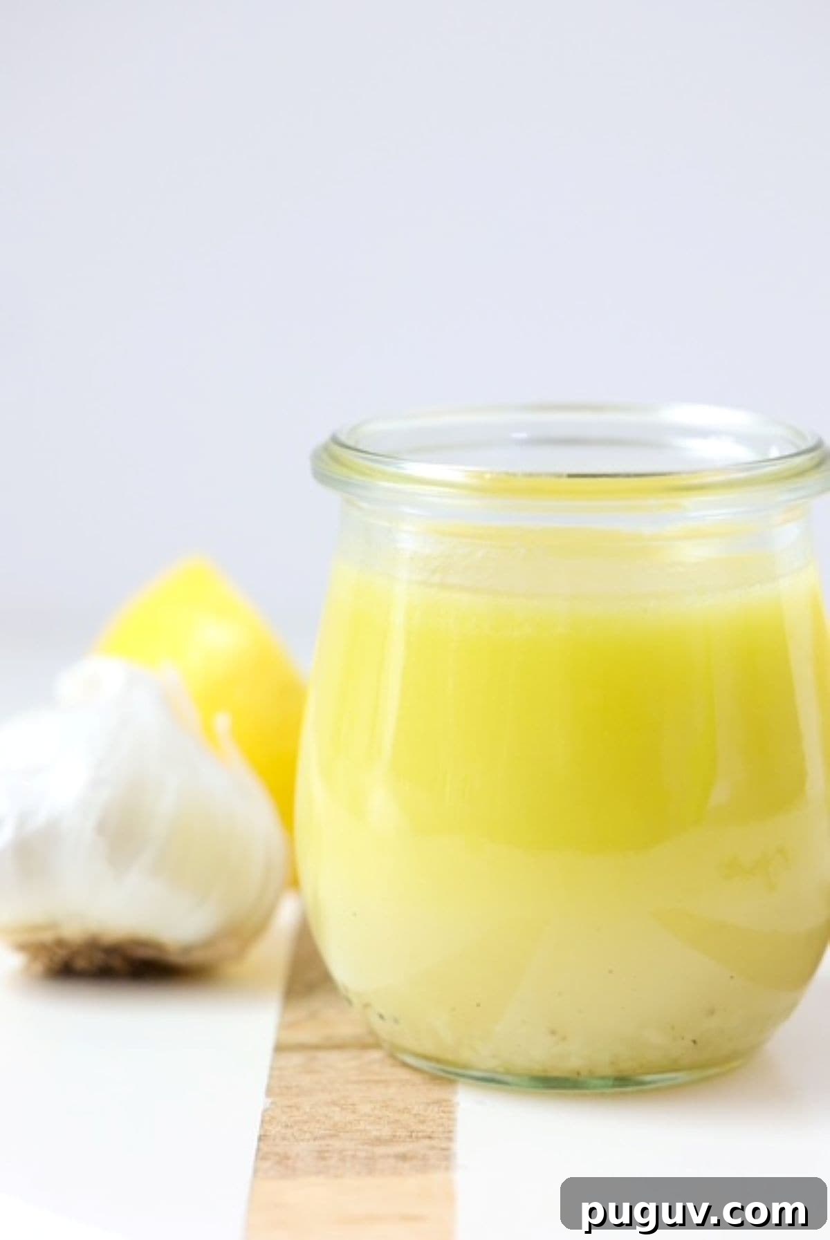 Lemon Garlic Salad Dressing Recipe: Bright Homemade Vinaigrette 2 Lemon Garlic Salad Dressing on a white board with a wooden stripe. One garlic bulb and lemon sit to the back right.