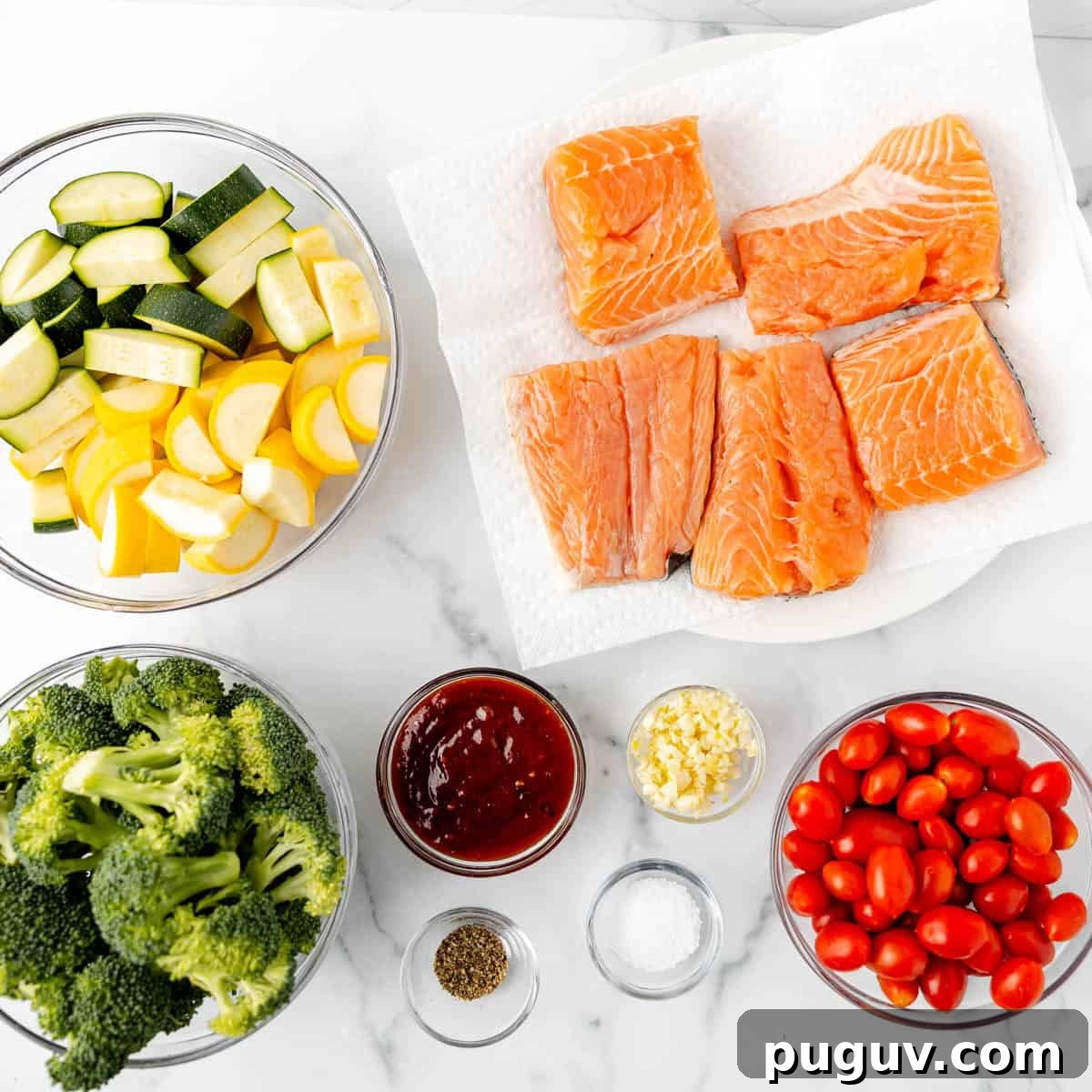 Bowls of zucchini, squash, raw salmon, broccoli, grape tomatoes, and small bowls of seasoning, garlic, and sauce on a white marble surface.