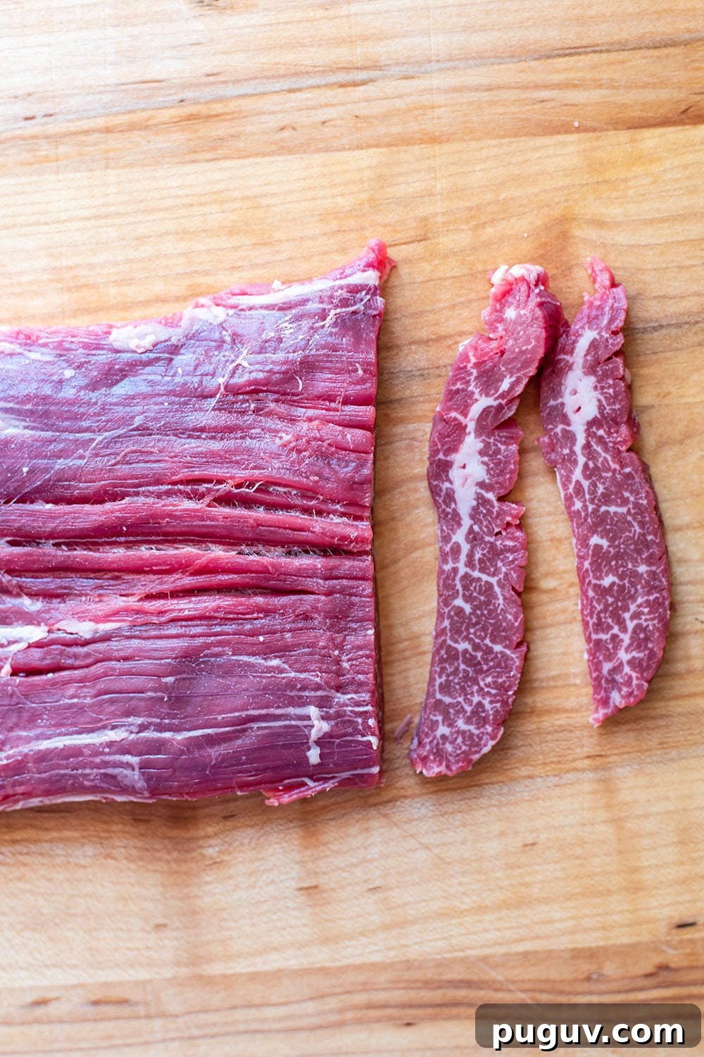 A raw uncooked flank steak on a wooden cutting board with two thin slices shaved off.
