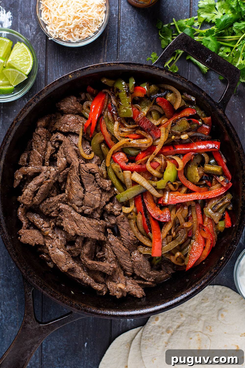 Skillet flank steak fajitas served in a cast iron skillet with bell peppers and onions alongside tortillas, cilantro, lime wedges, and shredded cheese.