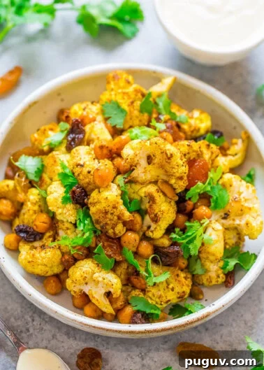 A bowl of roasted cauliflower and chickpeas garnished with herbs and served with a side of sauce.