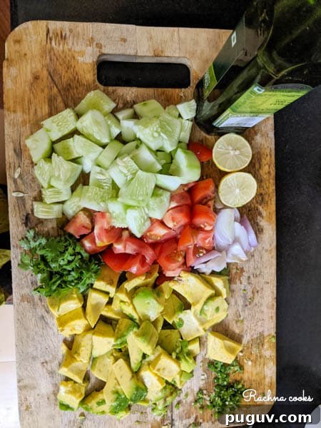 On a chopping board there are chopped cucumber, tomato, avocado, cilantro, onion, a lime and a bottle of olive oil.