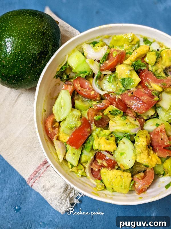 A close up of large avocado cucumber and tomato salad with onion slices in a white bowl with a blue background