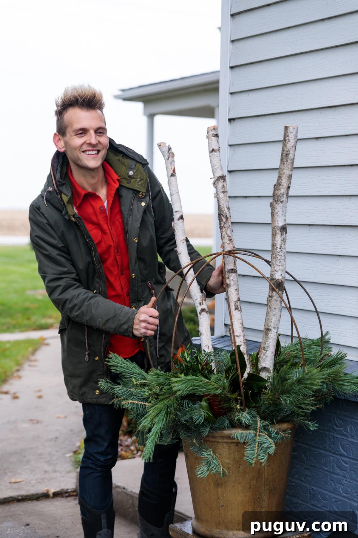 Kaleb Wyse wearing coat and red shirt holding birch sticks in a large urn with a round metal sphere with greens stuffed in amongst as well.