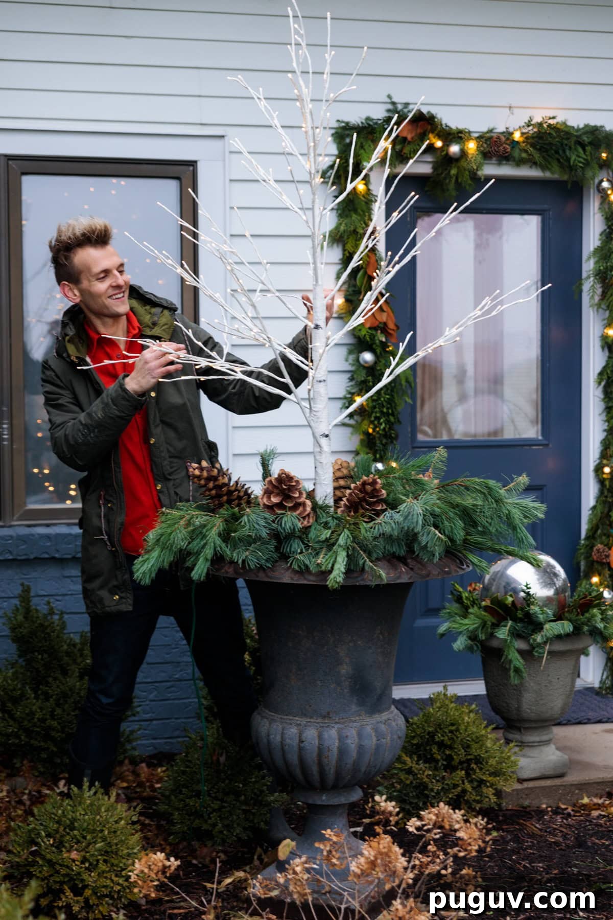 Kaleb Wyse wearing red shirt and coat placing a white tree with small lights on it into a large black urn with greens and pinecones all around the base.