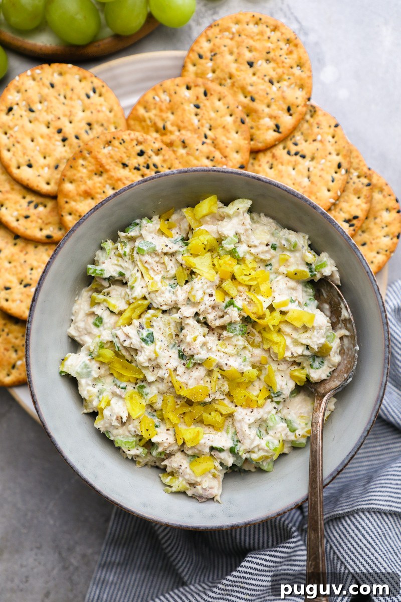 A serving bowl filled with pepperoncini tuna salad salad sitting next to wheat crackers and green grapes