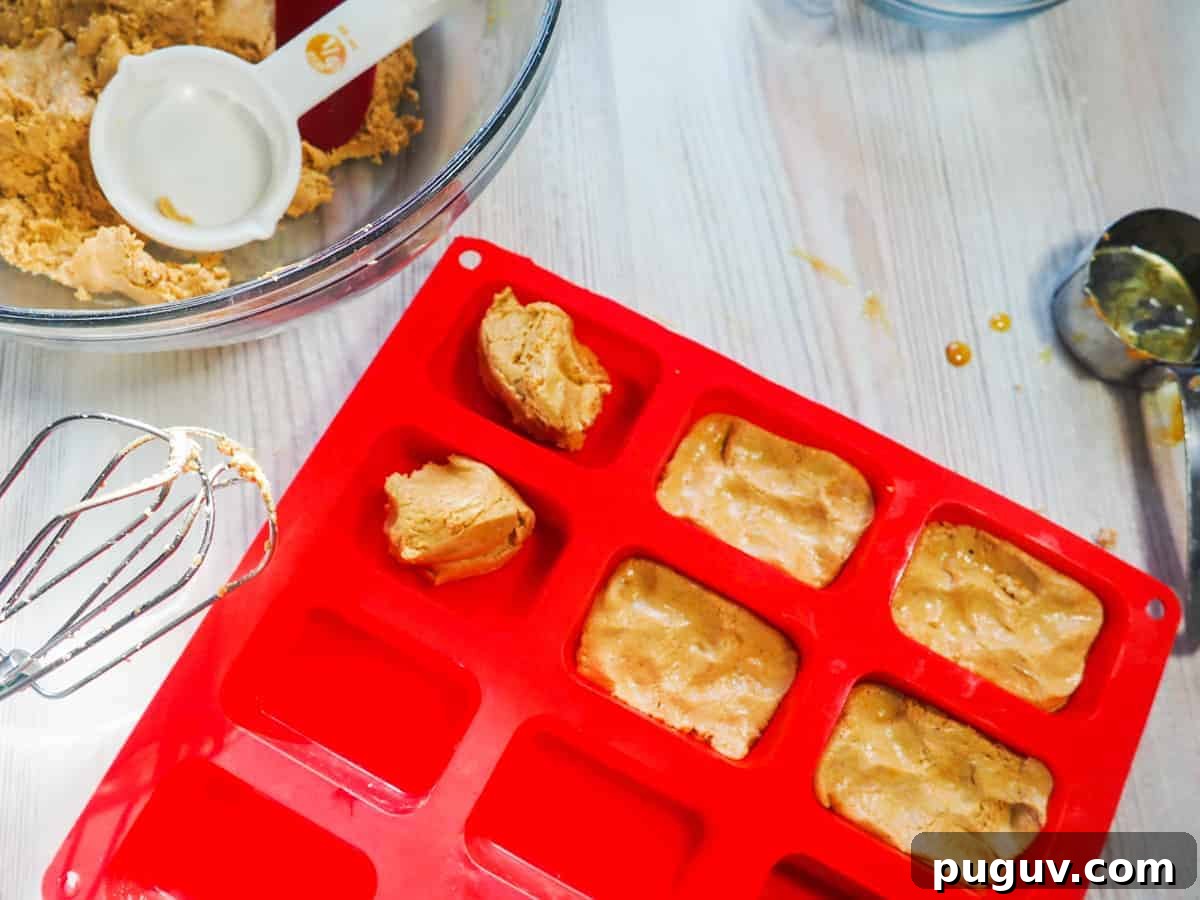 A red silicone loaf baking sheet being filled with peanut butter perfect bars and a mixing bowl with a red spatula and a measuring cup in it.