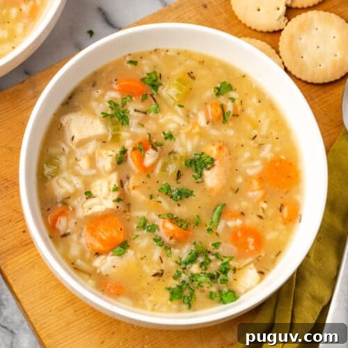 A bowl of turkey and rice soup sitting on a wood board next to some crackers.